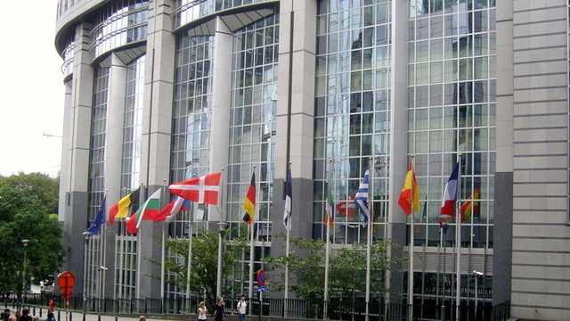 Exterior of the glass-and-concrete EU parliament building, ringed with flags.