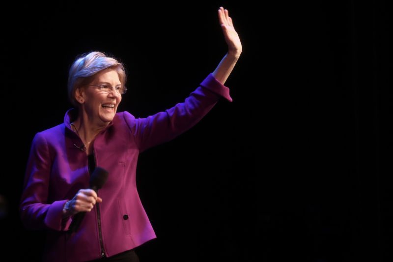 Elizabeth Warren holding a microphone and waving while speaking to a crowd.