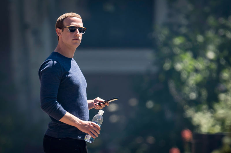 A man in sunglasses holds a cell phone and a water bottle while standing outside.