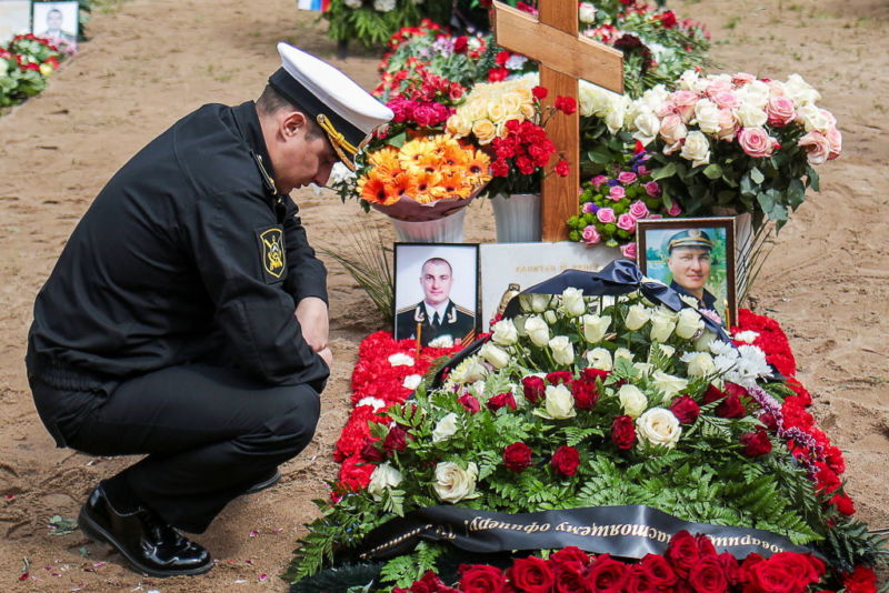 A man in uniform squats by a freshly dug grave.