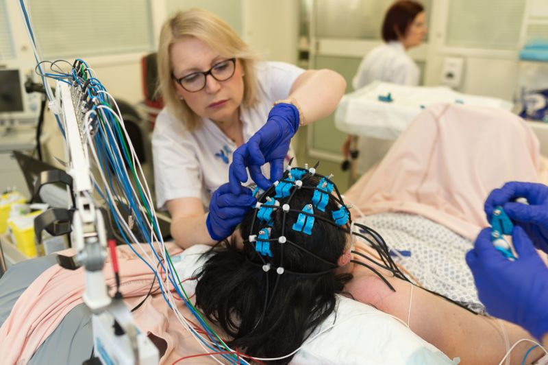 A woman in medical garb attaches electric devices to the head of a patient in bed.