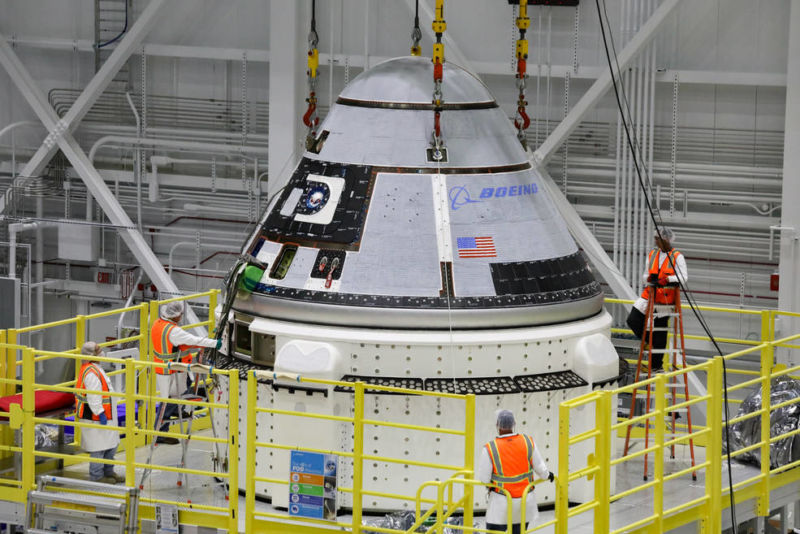 Orange-vested workers attend to a space capsule in a giant hangar.