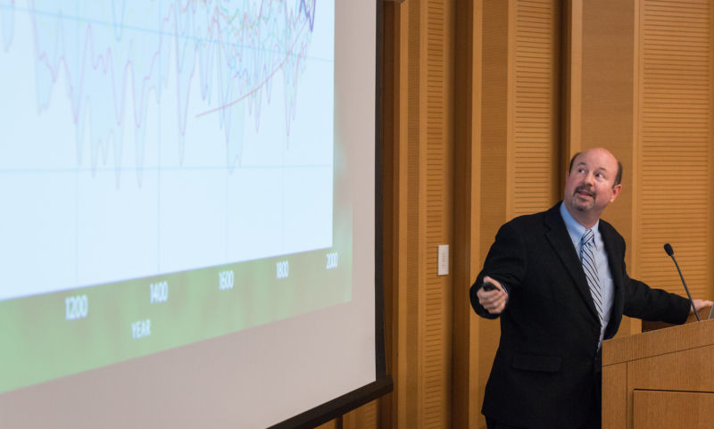 A man in a suit gestures toward a video projection.