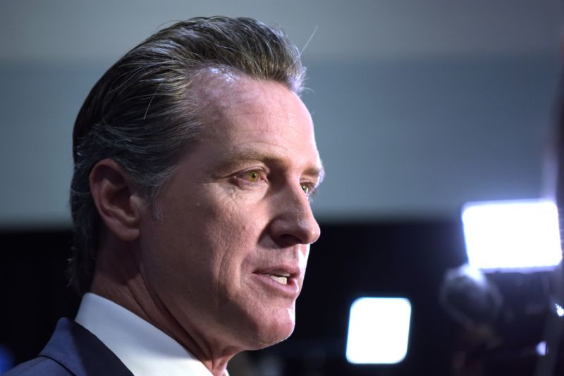 A close up of Newsom's face turned to face reporters not pictured, with camera lights in the background.