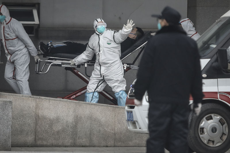 A person in full, white protective suit, blue face mask, and goggles, helps wheel a patient on a gurney into a hospital. His hand is outstretched as if he is signaling someone not to come near.