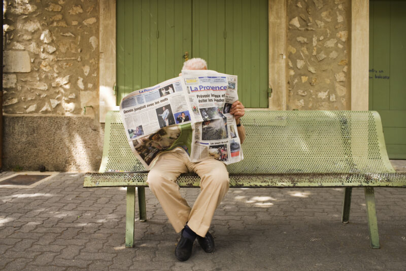 Photograph of a man sitting on a park bench reading a newspaper.