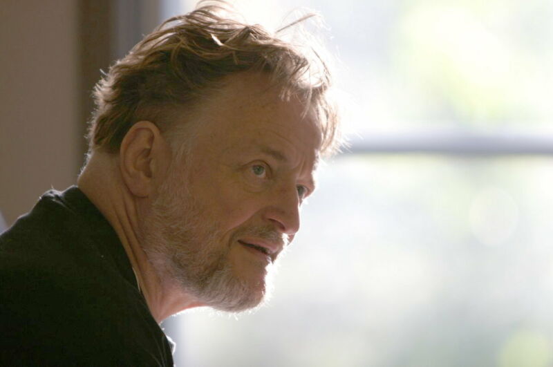 Photograph of a bearded man next to a window.