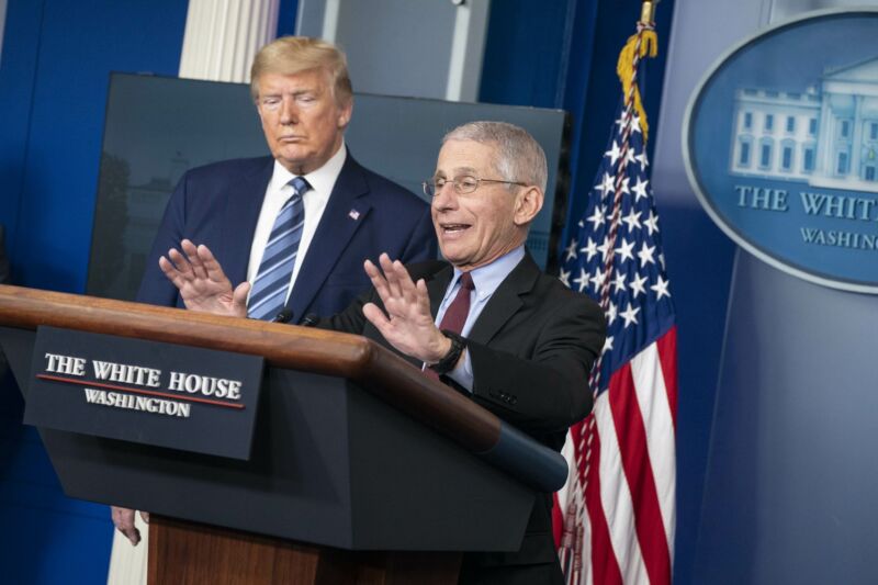 Anthony Fauci speaking at a podium during a press briefing while President Trump looks on.