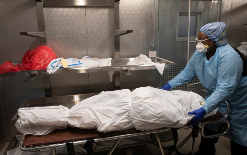 A medical technician in protective gear handles a wrapped corpse on a gurney.