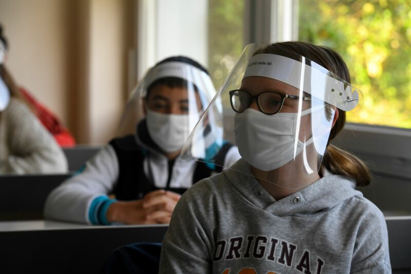 Children sit at desks while wearing clear masks.