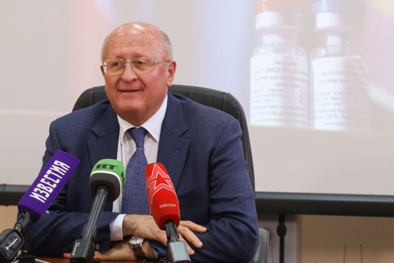 An older man in a suit speaks into an array of microphones.