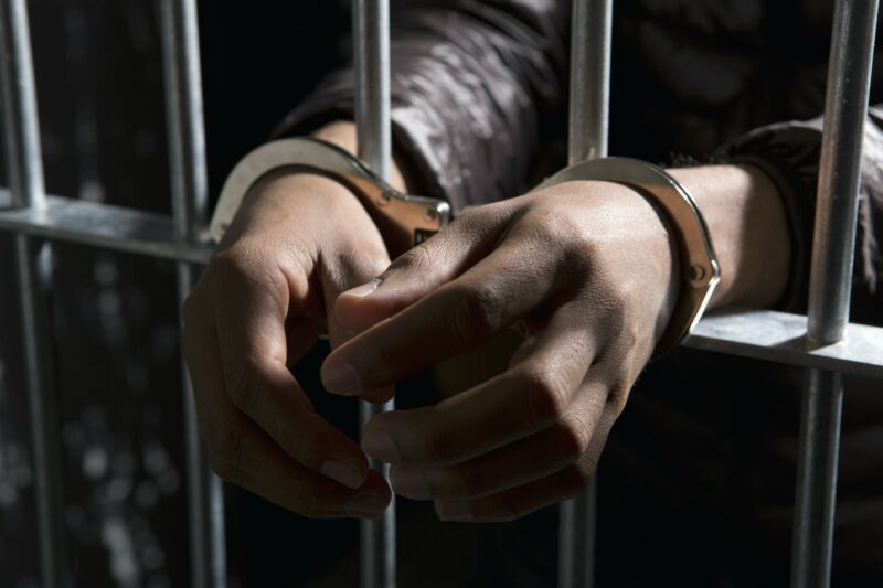 Picture of a jail cell in which a man's handcuffed hands are sticking out through the bars.