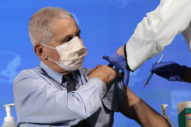 A masked man rolls up his sleeve to receive an injection.