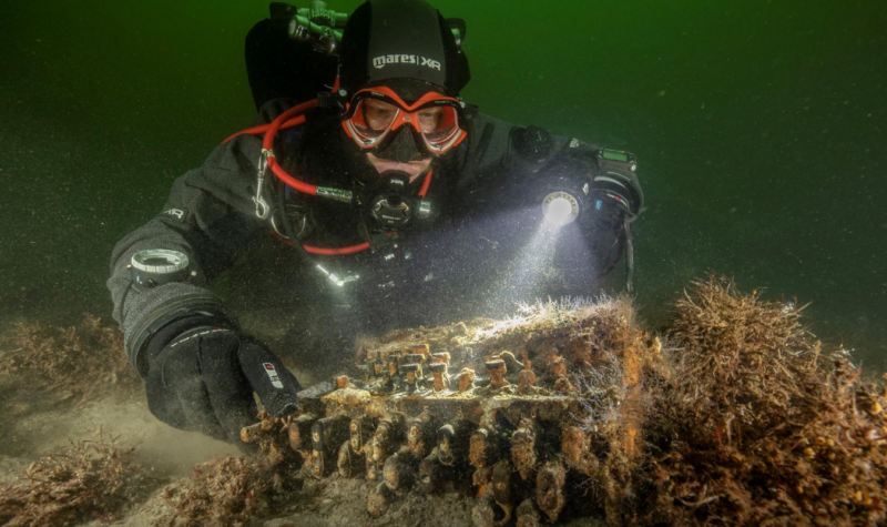 A deep-sea diver examines a heavily encrusted piece of machinery on the seabed.