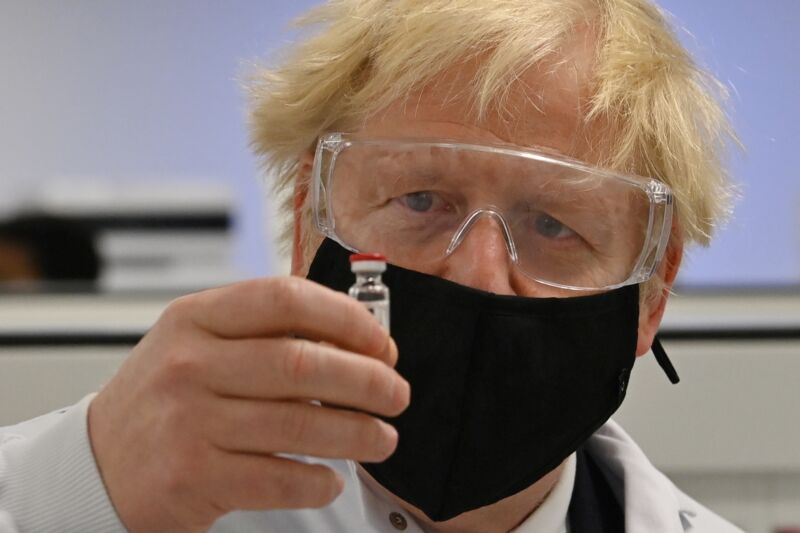 A man with a face mask, goggles, and tousled hair examines a small vial.