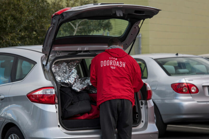 A man in a DoorDash-branded jacket reaches into the boot of a car.