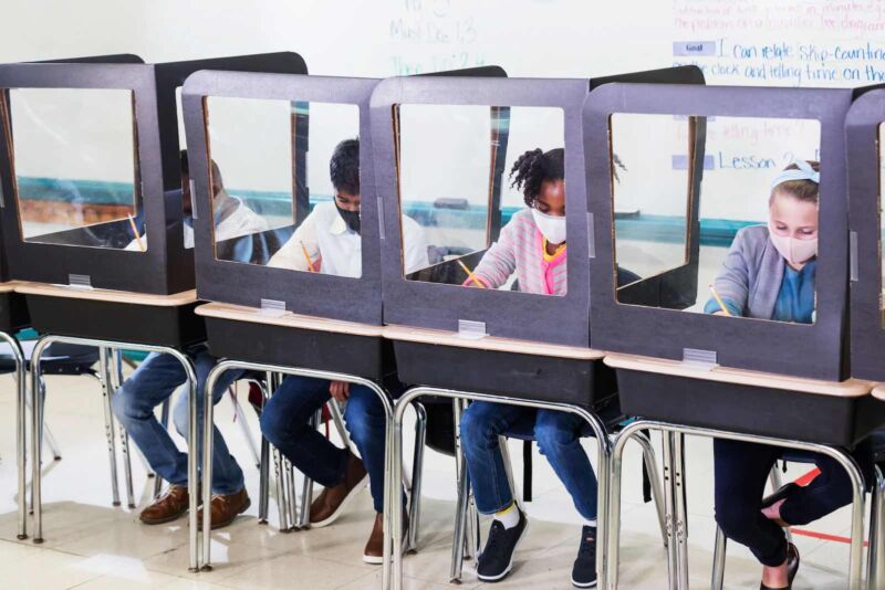 Masked school children work at desks separated by clear barriers.