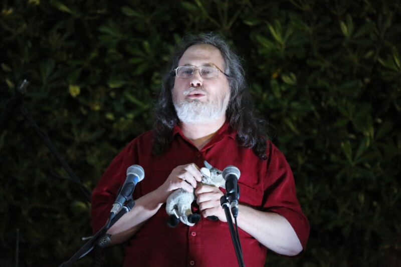 Richard Stallman speaking in front of microphones while holding a stuffed animal.