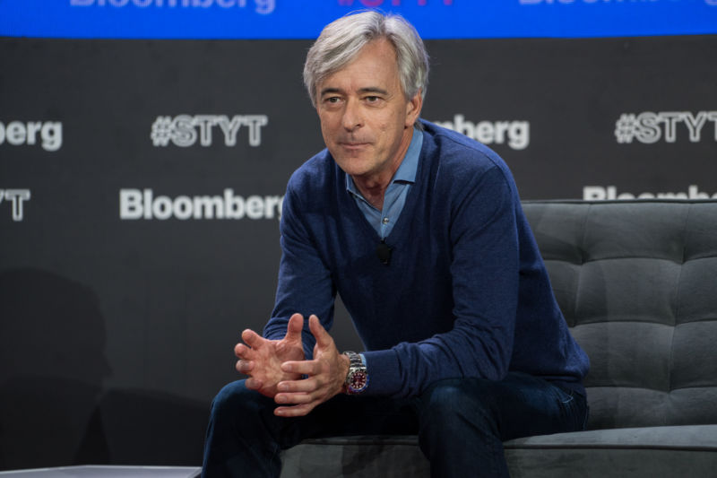 A middle-aged man sits on a stage during a presentation.