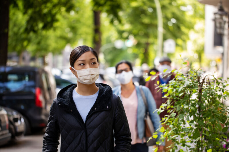 A masked woman walks along a treelined city street.