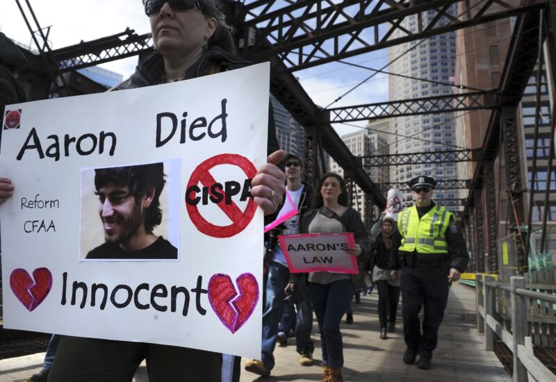 Protesters walking across a bridge. One holds a sign with a picture of Aaron Swartz that says, 