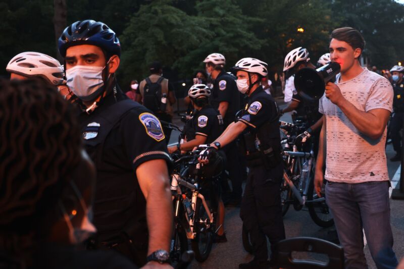 Police officers surround Jacob Wohl as he uses a megaphone to taunt anti-Trump protesters.