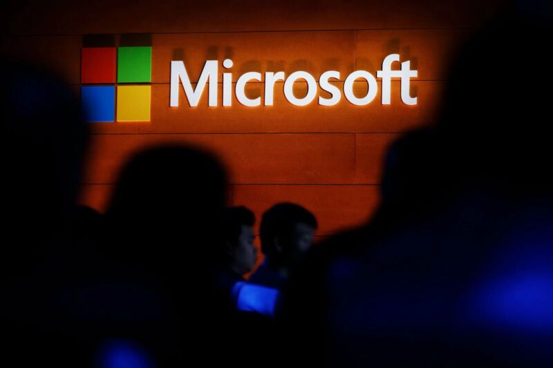 Shadowy figures stand beneath a Microsoft logo on a faux wood wall.