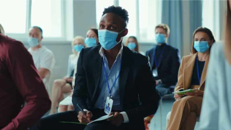 A man sits in the crowd, wearing a surgical mask, at an in-person event.