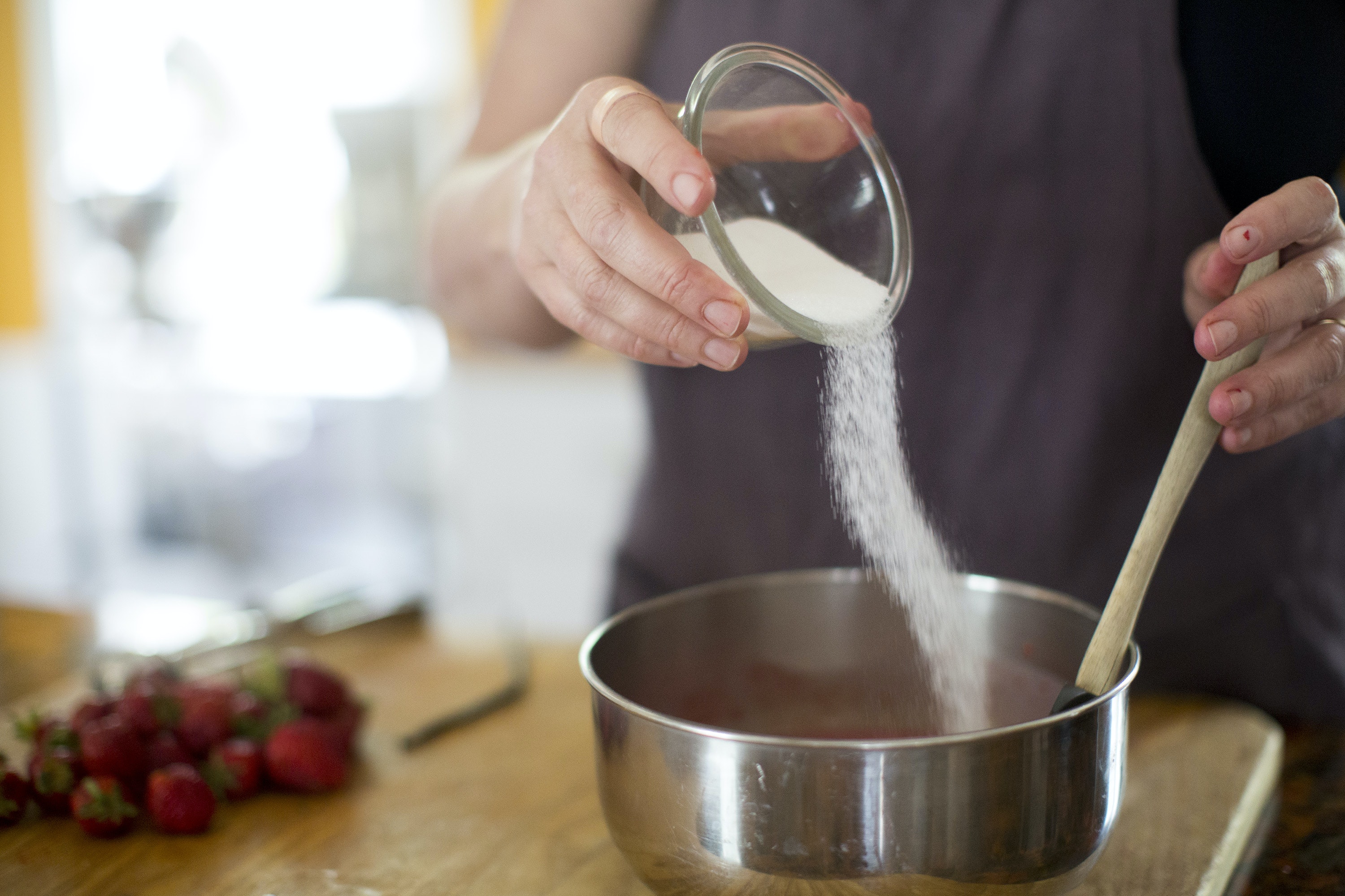 hands pouring sugar into a bowl with strawberries in the background