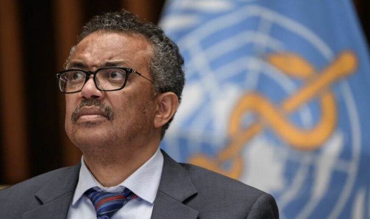 A man with a loosened necktie stands in front of a logo for the World Health Organization.