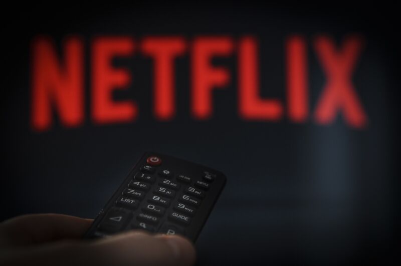 A person's hand holding a remote control in front of a TV screen with a Netflix logo.