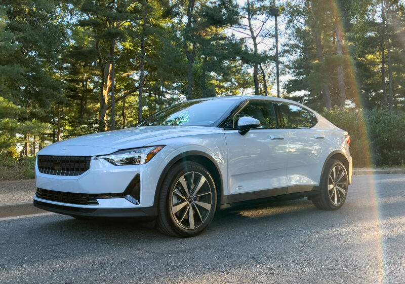 A white polestar 2 parked in the shade of some evergreens. The sun has hit the camera lens and created a rainbow next to the car