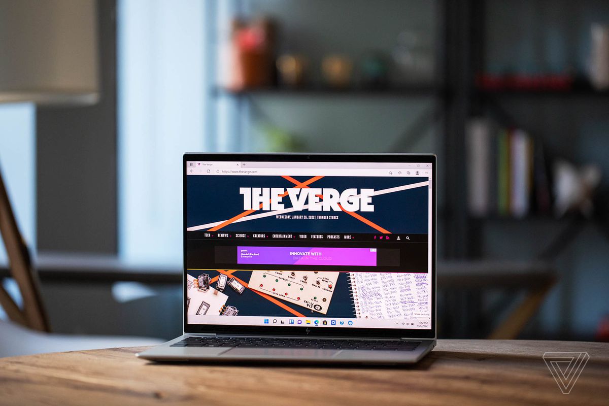 The HP Elite Dragonfly G3 on a wooden table with a bookshelf in the background, open, angled slightly to the left. The screen displays The Verge homepage.