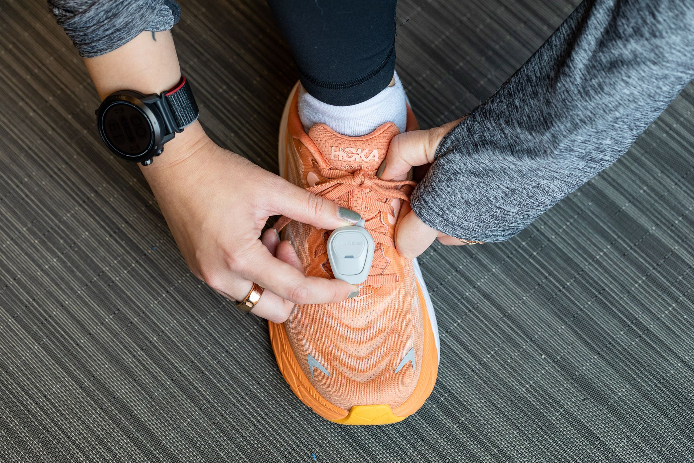 Coros Pod 2 on an orange Hoka shoe that’s being laced up by someone wearing a Coros Apex 2 Pro