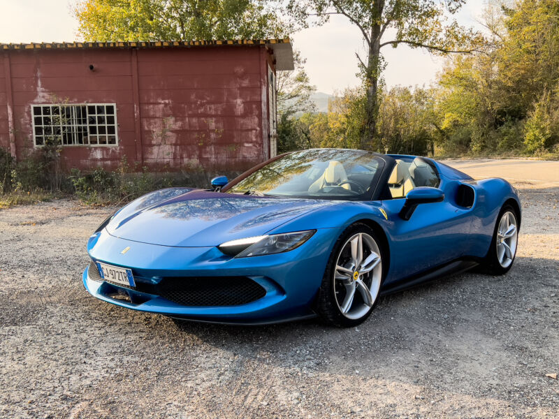 A blue Ferrari 296 GTS next to an old rural farm building