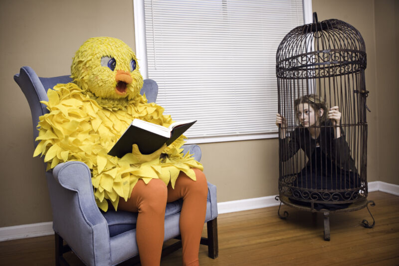 Person in full-size bird costume reads a book in a chair while a human looking distressed is locked in a human-sized bird cage.