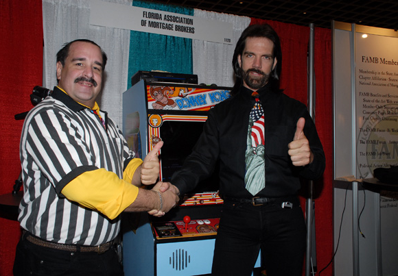 Mitchell (right) at the 2007 FAMB convention with former Twin Galaxies referee Todd Rogers and what appears to be a <em>Donkey Kong</em> cabinet with a modified joystick.”><figcaption class=