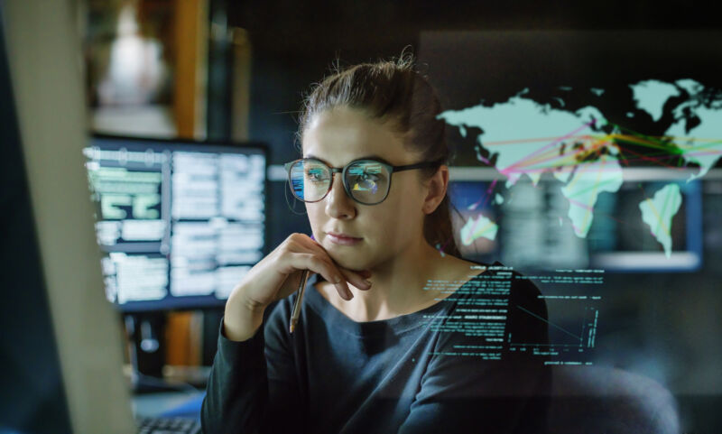 Stock image of a young woman, wearing glasses, surrounded by computer monitors in a dark office. In front of her there is a see-through displaying showing a map of the world with some data.