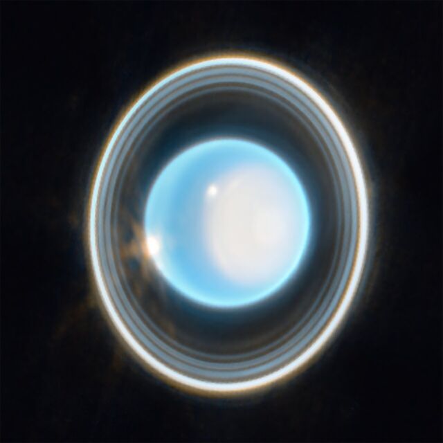 The planet Uranus on a black background. The planet appears light blue, with a large, white patch on the right side. On the edge of that patch at the upper left is a bright white spot. Another white spot is located on the left side of the planet at the 9 o’clock position. Around the planet is a system of nested rings. The rings of Uranus are vertical.