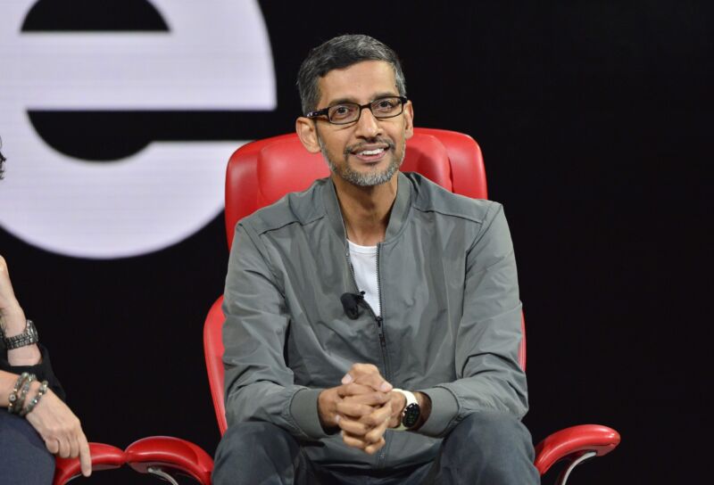 Google CEO Sundar Pichai smiling on stage at a conference where he was speaking. He sits in a comfortable chair and is leaning over with his hands clasped together.