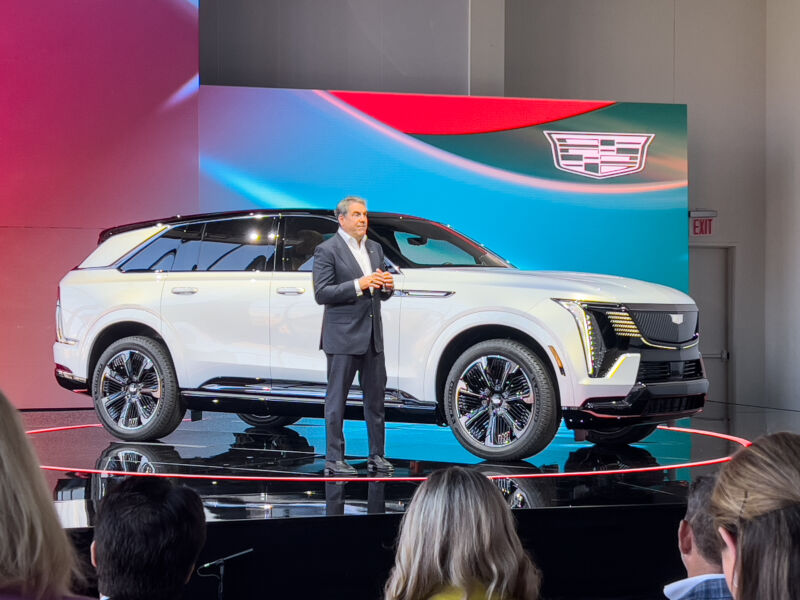 A man in a suit stands in front of a Cadillac Escalade IQ