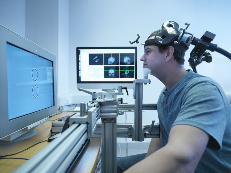 A person sits next to computer screens with other hardware strapped to his head.