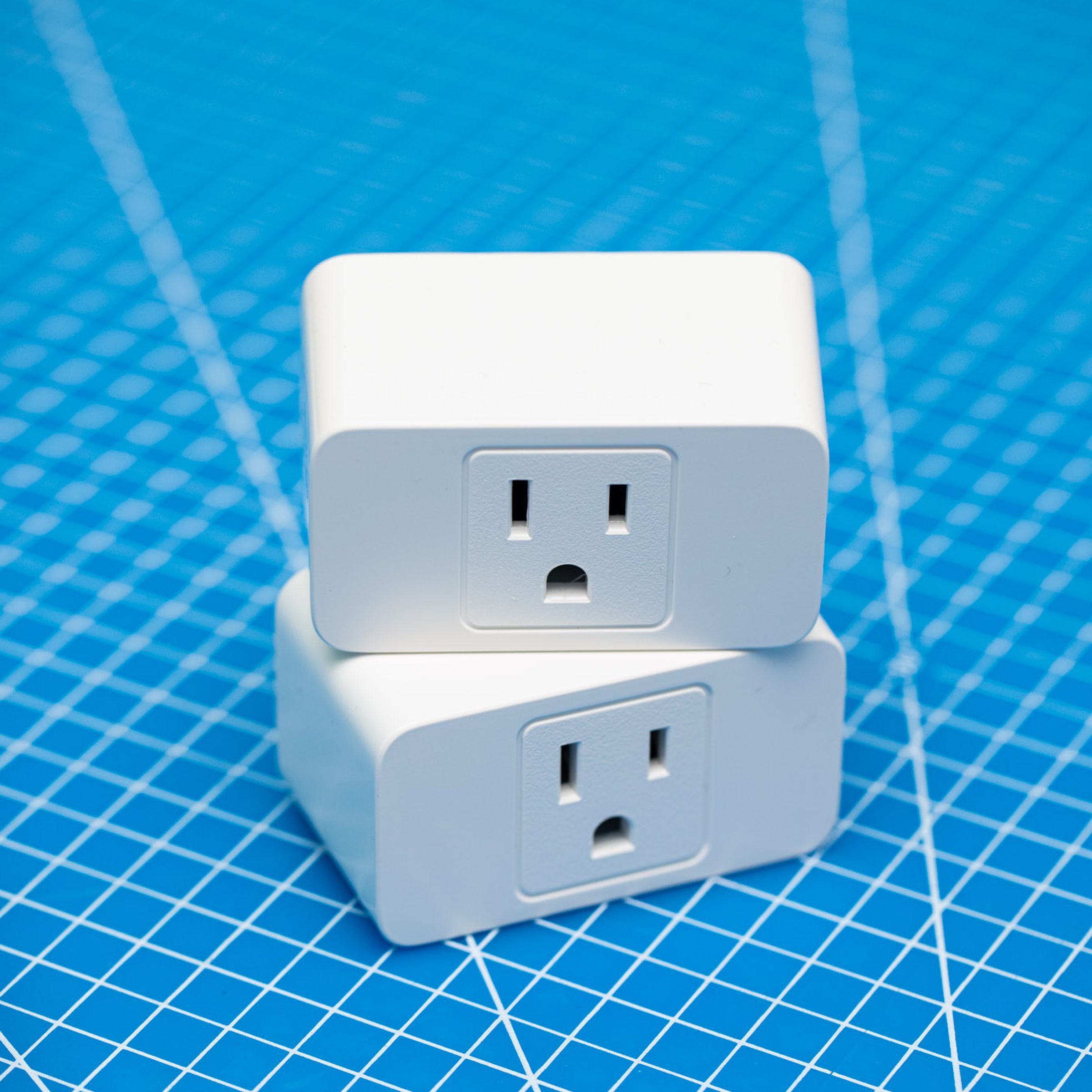 Two white Meross smart wi-fi plugs stacked on top of each other on a blue background. Each has a three-prong outlet on the front face.