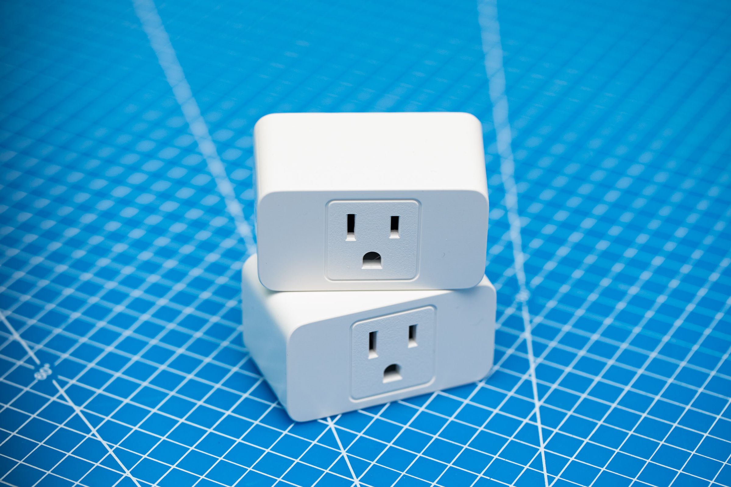 Two white Meross smart wi-fi plugs stacked on top of each other on a blue background. Each has a three-prong outlet on the front face.