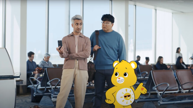 two men and a cartoon yellow bear standing in a departure area of an airport