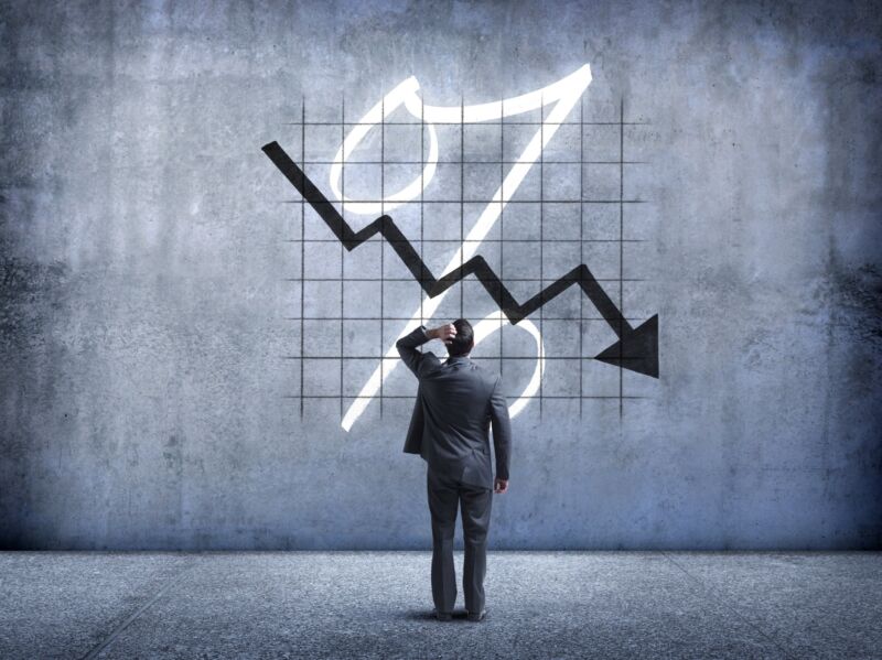 A businessman places his hand on his head as he looks up and is perplexed by a chart indicating a drop in value.