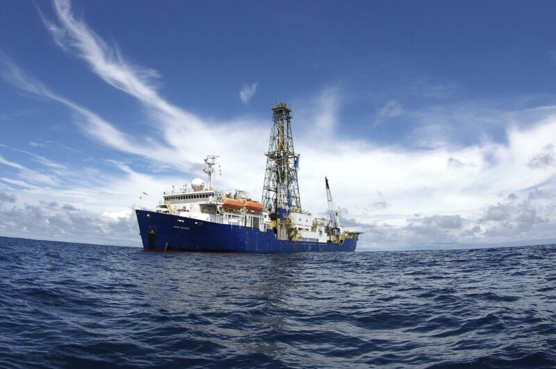 Image of a large boat with a tall tower at its center, and a crane in the rear. It is floating on a dark blue ocean and set in front of a white cloud.