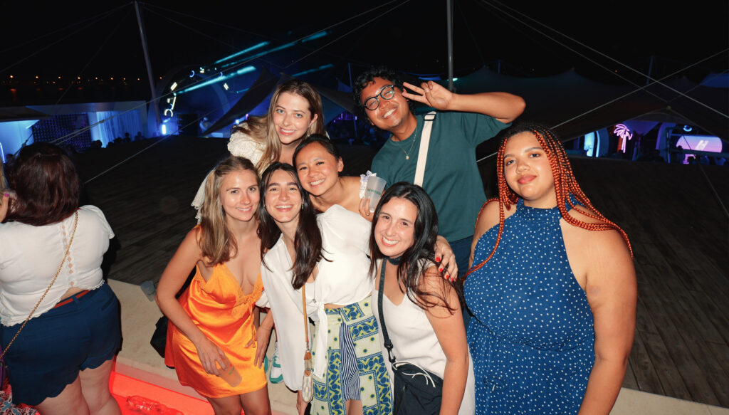 Group of young people on the beach at night smiling at the camera. Behind them is neon decor.