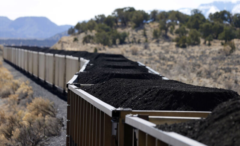 a long line of open-top rail cars filled with coal against a parched, scrub filled hill.