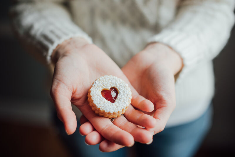 A woman in a white knit sweater, holding a Linzer cookie (with jam inside a heart cut-out) in her crossed palms.